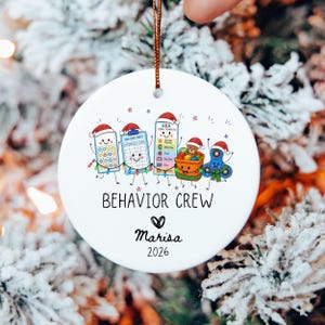 May include: A round white ceramic ornament featuring a colorful illustration of anthropomorphic therapy tools wearing Santa hats, including a pop-it, data sheet, schedule, toy bin, and fidget spinner. Text reads BEHAVIOR CREW, Marisa, and 2026.