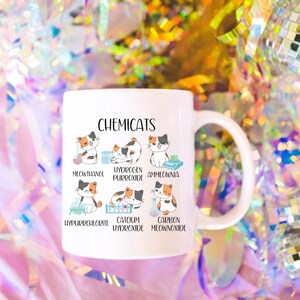 K&ouml;nnte beinhalten: Eine wei&szlig;e Keramiktasse mit einer Schwarzwei&szlig;illustration von Katzen mit chemischen Formeln. Der Text "CHEMICATS" befindet sich oben auf der Tasse. Die Katzenillustrationen sind mit den folgenden chemischen Formeln beschriftet: "MEOUTHANOL", "HYDROGEN PURROXIDE", "AMMEOWNIA", "HYPURRCHLORITE", "CATCIUM HYDROXIDE" und "CARBON MEOANNOXIDE".
