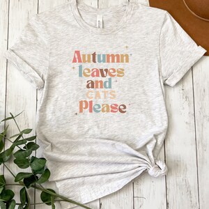 K&ouml;nnte beinhalten: Ein hellgraues T-Shirt mit dem Text "Autumn leaves and cats please" in bunten Buchstaben. Der Text ist in einem Retro-Schriftart-Stil.