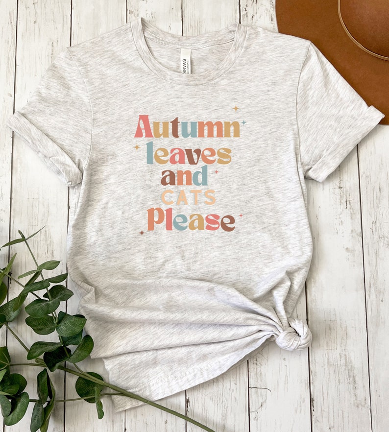 K&ouml;nnte beinhalten: Ein hellgraues T-Shirt mit dem Text "Autumn leaves and cats please" in bunten Buchstaben. Der Text ist in einem Retro-Schriftart-Stil.
