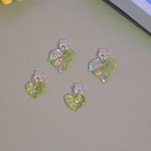 May include: Four leaf-shaped earrings with clear clips. The earrings are a light green color with iridescent highlights. The leaves have a textured appearance and are arranged on a light green surface.