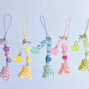 Puede incluir: Seis encantos de teléfono coloridos de Care Bear con cadenas de cuentas y detalles de estrellas y flores. Cada encanto presenta un Care Bear diferente con un símbolo único en su vientre.