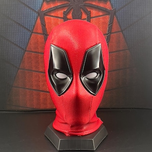 May include: A red Deadpool mask with black eye coverings and mesh eye inserts. The mask is displayed on a black stand, with a textured red fabric. The background features a spider web design.