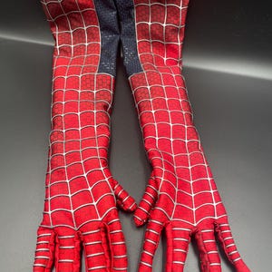 Op de afbeelding: Een paar rode en zilveren Spiderman-handschoenen met een webpatroon. De handschoenen zijn rood met een zilveren webdesign en hebben een donkerblauw gedeelte tussen de handschoenen.