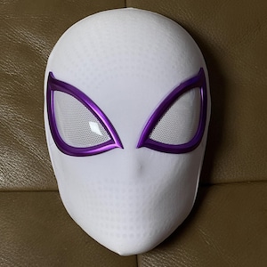 Op de afbeelding: Een wit Spider-Gwen-masker met een getextureerd oppervlak en paarse oogomlijsting. De oogopeningen zijn bedekt met een gaasmateriaal. Het masker is ontworpen om de vorm van een menselijk hoofd te volgen.