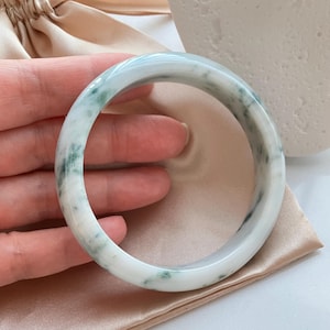 以下が含まれることがあります： 磨かれた石で作られた滑らかな円形のバングルブレスレット。素材は主に半透明の白で、バンドの表面全体に深緑色の不規則な斑点がはっきりと散らばっているのが特徴です。