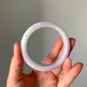 May include: A translucent jade bangle bracelet with a gradient of white and lavender hues. The smooth, polished surface reflects light, showcasing the natural variations in color. This elegant piece of jewelry is a classic accessory.