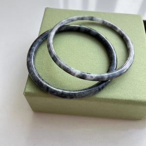 Puede incluir: Dos brazaletes circulares con un patrón jaspeado en tonos gris carbón, negro y blanco. Las pulseras tienen un acabado liso y pulido con un perfil redondeado, mostrando una textura continua y arremolinada en cada una de las piezas.