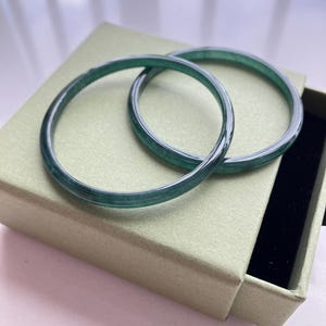 May include: Two smooth, circular dark green bangles with a translucent, marbled appearance. The bracelets feature a polished finish and are resting on a light olive green square box with a black interior lining, showcasing a simple, rounded design.