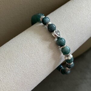 May include: A beaded bracelet featuring round, dark green mottled stone beads interspersed with silver-toned metal spacers and a single heart-shaped charm with a textured, dotted surface pattern, strung on an elastic cord.