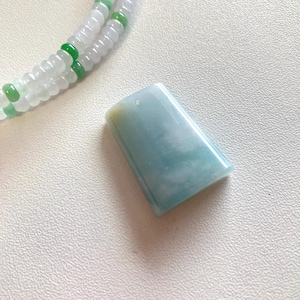 May include: A rectangular, pale blue jade pendant with a subtle, cloudy pattern. A small hole is drilled at the top. A beaded necklace with white and green beads is in the background. The pendant is likely for a necklace or bracelet.