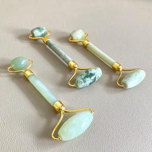Op de afbeelding: Drie jade gezichtsrollers met goudkleurige metalen accenten. Elke roller heeft een goudkleurig frame met een gladde, ovaalvormige jade steen en een kleinere, afgeronde jade steen. De jadestenen variëren in kleur van lichtgroen tot een gevlekt groen en wit.