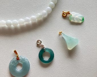 May include: An overhead shot showcases a collection of jade jewelry. A white beaded necklace is at the top. Below are three jade pendants in shades of green and blue, and a circular jade pendant. The pendants have gold and silver accents.