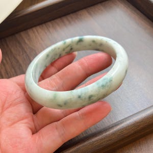 May include: A smooth, polished bangle bracelet crafted from light green and white stone with mottled dark green inclusions throughout the circular band. The jewelry piece features a rounded profile and a glossy, reflective surface finish.