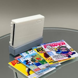 Puede incluir: Una consola Nintendo Wii blanca con una base gris, rodeada de varias copias de la revista Nintendo Power con un personaje de dibujos animados en la portada. La portada de la revista dice "Nintendo Power" y "Ready to Ware? Donkey Kong Country Returns".