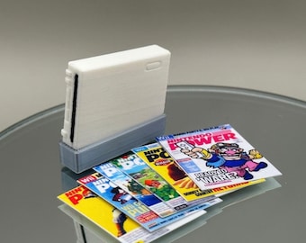 Puede incluir: Una consola Nintendo Wii blanca con una base gris, rodeada de varias copias de la revista Nintendo Power con un personaje de dibujos animados en la portada. La portada de la revista dice "Nintendo Power" y "Ready to Ware? Donkey Kong Country Returns".