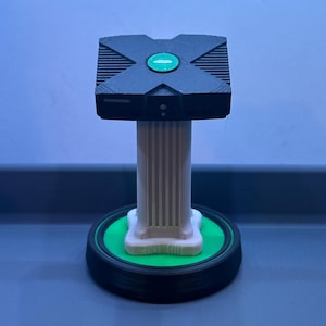 May include: A black and green decorative stand featuring a miniature replica of a classic video game console. The console sits atop a white column with a green base and a black circular platform. The stand is likely 3D-printed.