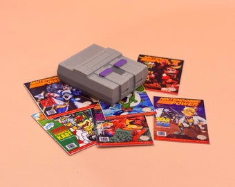 Puede incluir: Una consola Super Nintendo Entertainment System (SNES) gris con una ranura de cartucho púrpura, rodeada de varias revistas Nintendo Power con diferentes portadas de videojuegos, incluyendo 'Mega Man X', 'Mario Kart', 'Star Fox' y 'Yoshi's Island'.