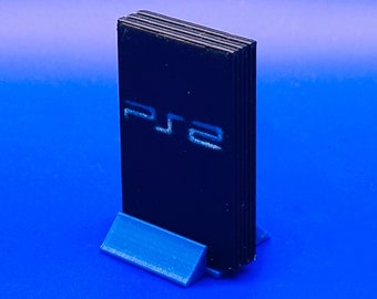 Puede incluir: Una consola PlayStation 2 negra en un soporte azul. La consola tiene el logotipo de PlayStation 2 en la parte delantera.
