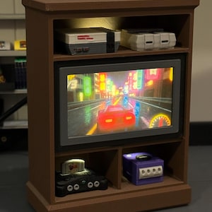 Könnte beinhalten: Ein Miniatur-Unterhaltungsmöbel aus braunem Holz mit einem eingebauten Fernseher, der ein Rennspiel zeigt. Die Regale beherbergen Miniatur-Spielkonsolen, darunter ein Nintendo Entertainment System, Super Nintendo, Nintendo 64 und GameCube.