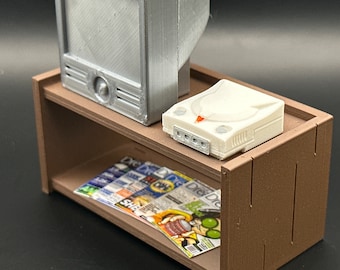 Puede incluir: Una estantería de madera en miniatura con una consola Sega Dreamcast blanca en la repisa superior, un televisor plateado en la repisa inferior y una revista en la repisa inferior.