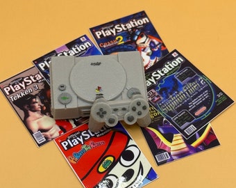 Puede incluir: Una consola PlayStation gris con un mando gris, rodeada de varias portadas de revistas PlayStation con personajes de videojuegos populares como Crash Bandicoot, Tekken y Spyro the Dragon.