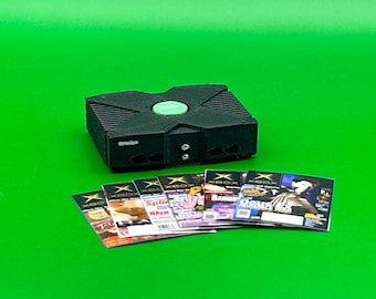 Puede incluir: Una consola Xbox negra con un botón verde en la parte superior y seis revistas de videojuegos con la palabra "Xbox" en la portada.
