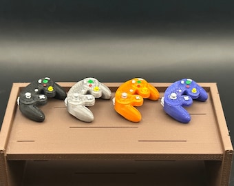 May include: Four miniature GameCube controllers in different colours: black, silver, orange, and purple. The controllers are arranged in a row on a brown wooden surface.