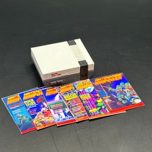 Pode incluir: Um console Nintendo Entertainment System (NES) branco e cinza com seis revistas Nintendo Power. As revistas apresentam capas com personagens de Duck Tales, Rescue Rangers, Battletoads e Teenage Mutant Ninja Turtles.