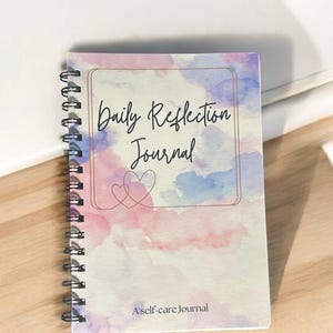 May include: A spiral bound journal with a watercolor design in shades of pink, blue, and purple. The cover features the text "Daily Reflection Journal" and "A self-care journal".