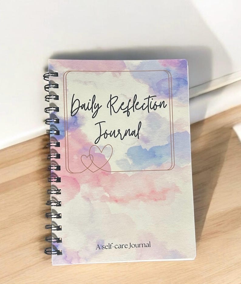 May include: A spiral bound journal with a watercolor design in shades of pink, blue, and purple. The cover features the text "Daily Reflection Journal" and "A self-care journal".