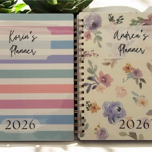 Może przedstawiać: Dwa planery w oprawie spiralnej. Jeden ma wz&oacute;r w paski w pastelowych kolorach z napisem "Korin's Planner" i "2026". Drugi ma kwiatowy wz&oacute;r z napisem "Andrea's Planner" i "2026".