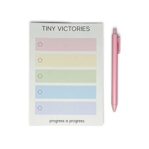 May include: A rectangular notepad with the text TINY VICTORIES at the top and progress is progress. at the bottom. It features five horizontal pastel-colored rows, each with a star icon on the left. A solid light pink retractable pen is included.