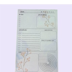 May include: A light blue daily planner with floral accents. The planner includes sections for date, to-do list, schedule, notes, priorities, appointments, and a gratitude section. The design features a watercolor floral motif.