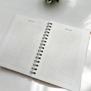 May include: An open spiral bound journal with lined pages. The journal is white with black text that says "Daily Journal" on both pages.