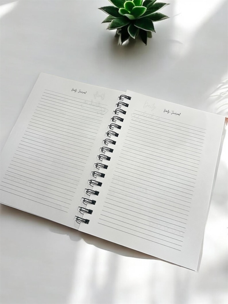 May include: An open spiral bound journal with lined pages. The journal is white with black text that says "Daily Journal" on both pages.