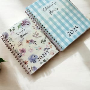 Może przedstawiać: Dwa spiralnie oprawione planery. Jeden ma jasnoniebieski wz&oacute;r w kratkę vichy z napisem "Adrianna's Planner" i "2025". Drugi ma kwiatowy wz&oacute;r z napisem "Laura's Planner" i "2025". Oba są otwarte.