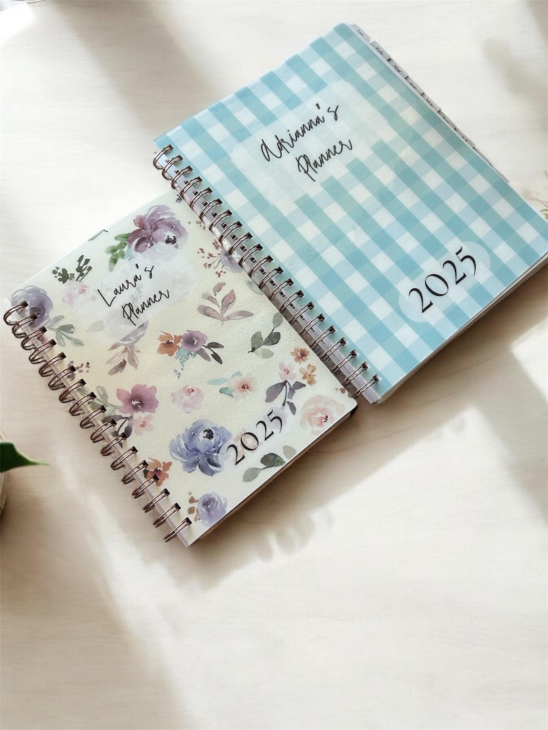 Może przedstawiać: Dwa spiralnie oprawione planery. Jeden ma jasnoniebieski wz&oacute;r w kratkę vichy z napisem "Adrianna's Planner" i "2025". Drugi ma kwiatowy wz&oacute;r z napisem "Laura's Planner" i "2025". Oba są otwarte.
