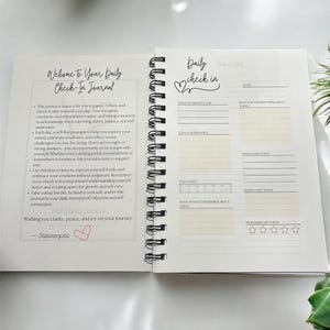 May include: A white journal with a black spiral binding. The cover features the text "Welcome to Your Daily Check-In Journal" with a pink heart. The inside pages have a daily check-in section with prompts for gratitude, affirmations, feelings, and a rating of the day.