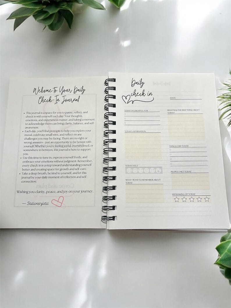 May include: A white journal with a black spiral binding. The cover features the text "Welcome to Your Daily Check-In Journal" with a pink heart. The inside pages have a daily check-in section with prompts for gratitude, affirmations, feelings, and a rating of the day.