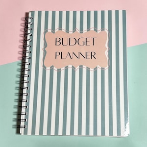 May include: A spiral-bound budget planner featuring a vertical striped pattern in white and muted blue. Centered on the cover is a light pink scalloped-edge label with the black text BUDGET PLANNER printed in a clean, sans-serif typeface.