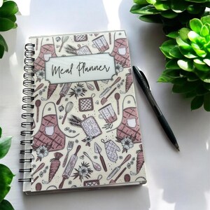 May include: A spiral-bound meal planner notebook with a cream-colored cover featuring a repeating pattern of kitchen utensils, aprons, and flowers in shades of brown and pink, with the text Meal Planner centered in a rectangular frame.