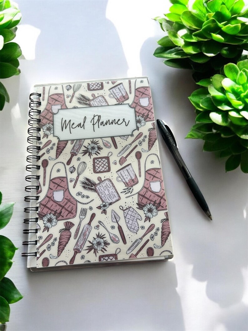 May include: A spiral-bound meal planner notebook with a cream-colored cover featuring a repeating pattern of kitchen utensils, aprons, and flowers in shades of brown and pink, with the text Meal Planner centered in a rectangular frame.
