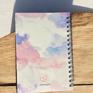 May include: A spiral bound notebook with a watercolor design of pink and blue clouds. The back cover has a logo with the text "Stationeryistic" and a paperclip heart.