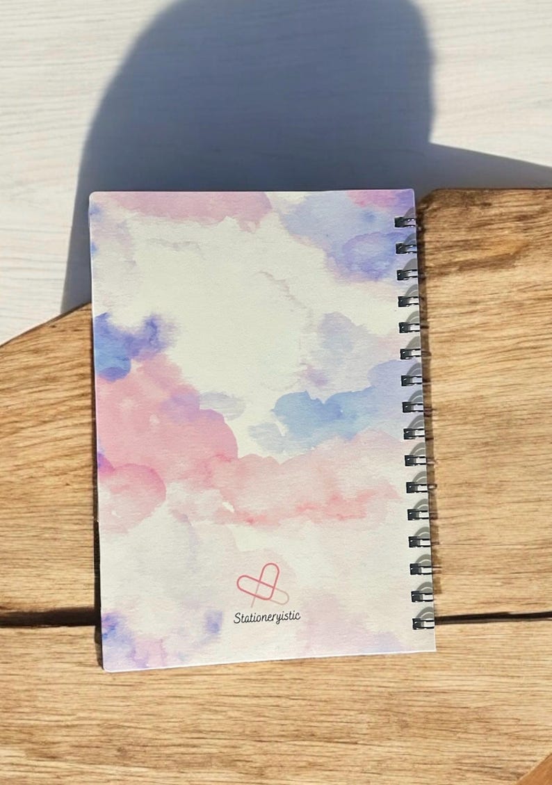 May include: A spiral bound notebook with a watercolor design of pink and blue clouds. The back cover has a logo with the text "Stationeryistic" and a paperclip heart.