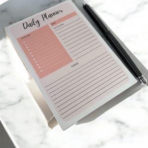 May include: A daily planner notepad with a pink and white design. The notepad includes sections for a to-do list, date, top priorities, and notes. A black pen rests on the notepad.