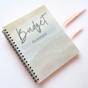 May include: A spiral-bound budget planner with a beige and white watercolor-style cover. The front cover features the word Budget in a large script font and the word PLANNER in a smaller sans-serif font. Two light pink tassels extend from the side.