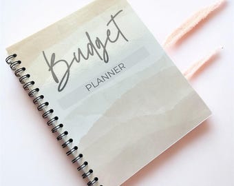May include: A spiral-bound budget planner with a watercolor-style cover. The cover features the word "Budget" in cursive script above the word "PLANNER". The color palette includes beige, light blue, and white. Two pink, feathery accents are visible.