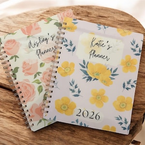 Może przedstawiać: Dwa spiralnie oprawione planery z kwiatowymi wzorami. Jeden ma r&oacute;żowe r&oacute;że, drugi ż&oacute;łte kwiaty oraz tekst "Katie's Planner" i "2026". Leżą na drewnianej powierzchni.