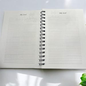 May include: An open spiral bound notebook with lined pages. The notebook is white with black text that says "Daily Journal" on both sides.